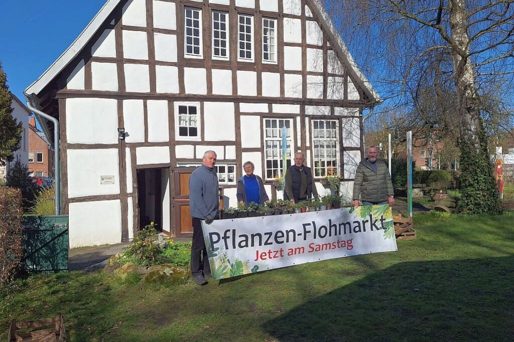 Pflanzenflohmarkt 2026 22 Vier Personen stehen vor einem Fachwerkhaus. Davor steht ein Schild mit dem Text Pflanzen-Flohmarkt, Jetzt am Samstag.