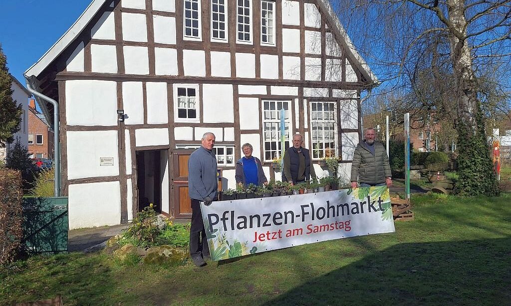 Vier Personen stehen vor einem Fachwerkhaus. Davor steht ein Schild mit dem Text Pflanzen-Flohmarkt, Jetzt am Samstag.