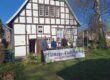 Vier Personen stehen vor einem Fachwerkhaus. Davor steht ein Schild mit dem Text Pflanzen-Flohmarkt, Jetzt am Samstag.