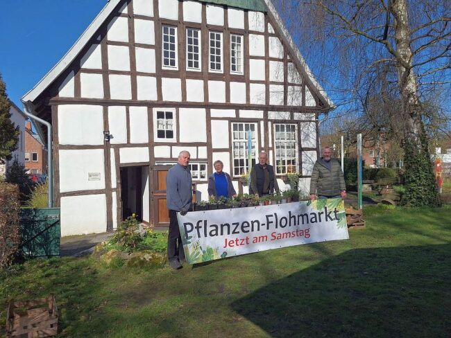 Vier Personen stehen vor einem Fachwerkhaus. Davor steht ein Schild mit dem Text Pflanzen-Flohmarkt, Jetzt am Samstag.