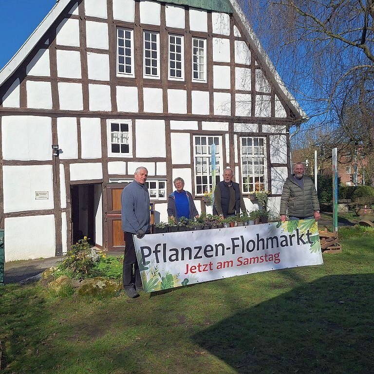 Vier Personen stehen vor einem Fachwerkhaus. Davor steht ein Schild mit dem Text Pflanzen-Flohmarkt, Jetzt am Samstag.