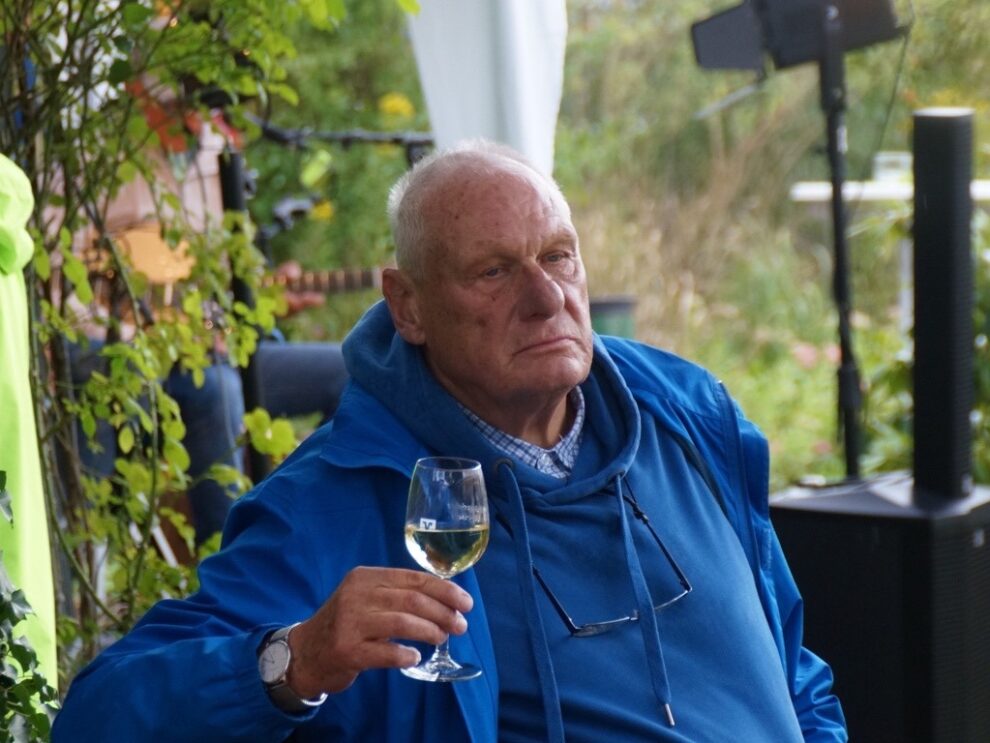 Ein Mann in blauer Jacke sitzt im Freien und hält ein Glas Weißwein.