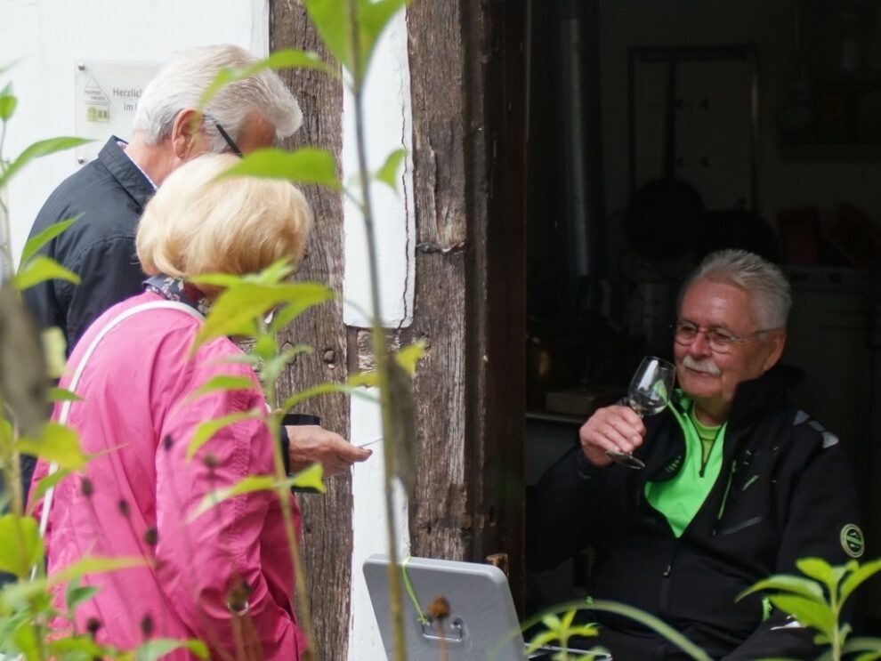 Ein sitzender Mann hebt sein Weinglas im Gespräch mit zwei Gästen an einer Tür.