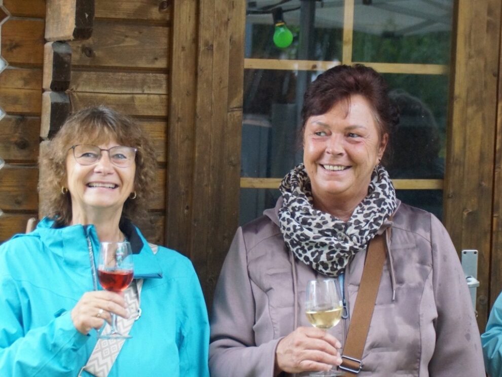 Zwei Frauen lächeln mit gefüllten Weingläsern vor einer Holzhütte.