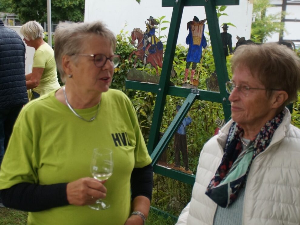 Eine Helferin und eine Besucherin unterhalten sich mit Weinglas vor einer bunten Dekoration.
