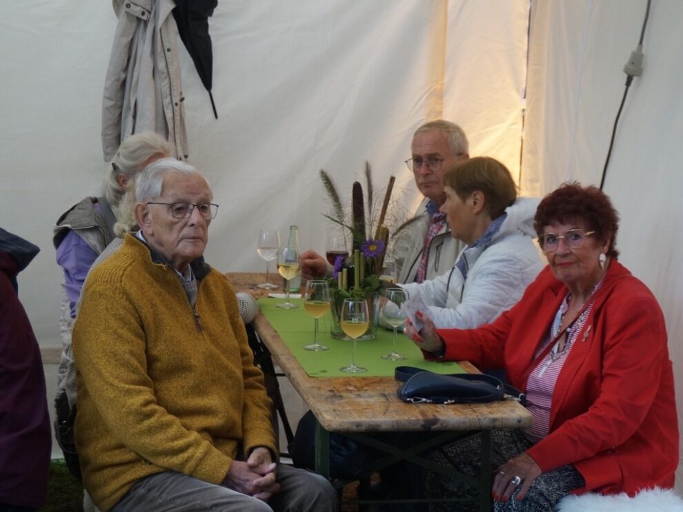 Mehrere ältere Gäste sitzen mit Wein und Wasser gemeinsam an einer langen Tafel.