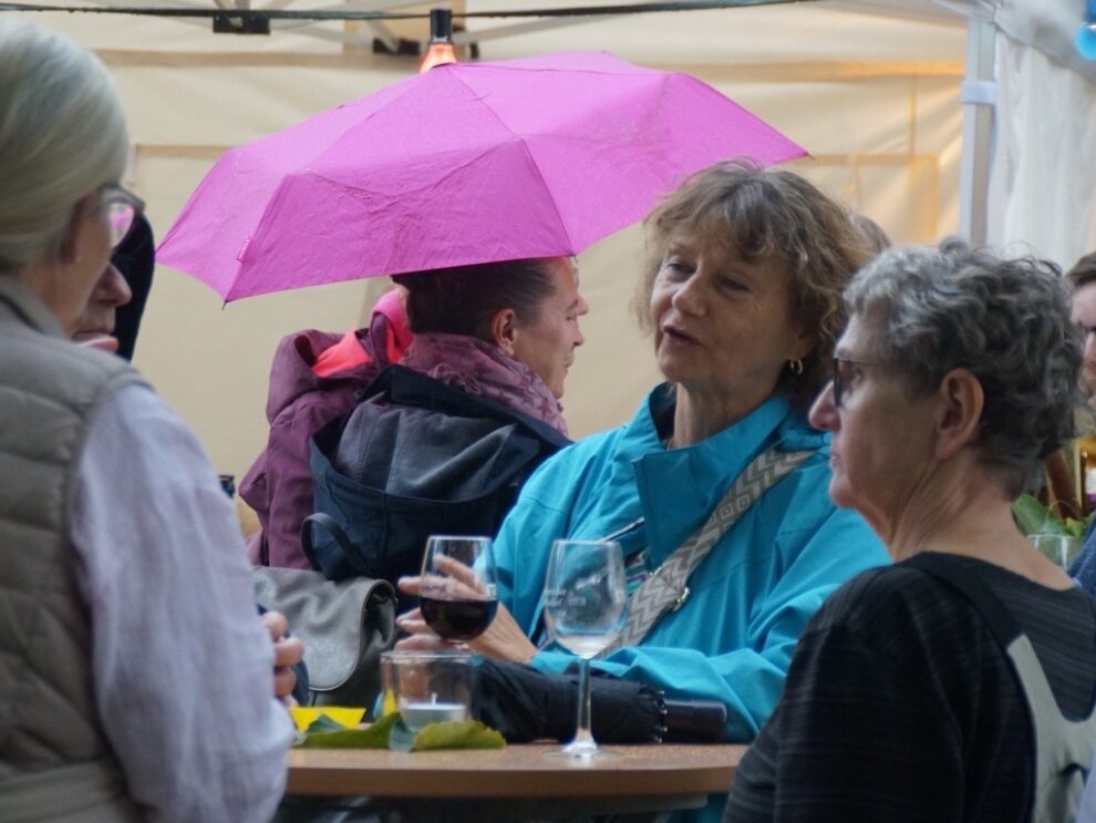 Mehrere Frauen unterhalten sich bei Regen mit Weingläsern unter einem pinkfarbenen Schirm.