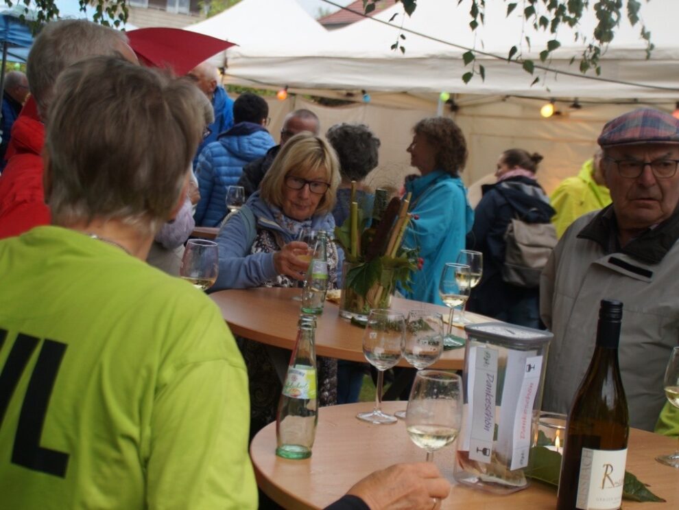 Gäste und Helferinnen stehen mit Weingläsern an geschmückten Tischen im Festzelt.