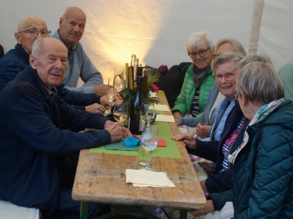 Eine größere Gruppe älterer Gäste sitzt mit Wein und Wasser an einer langen Tafel.