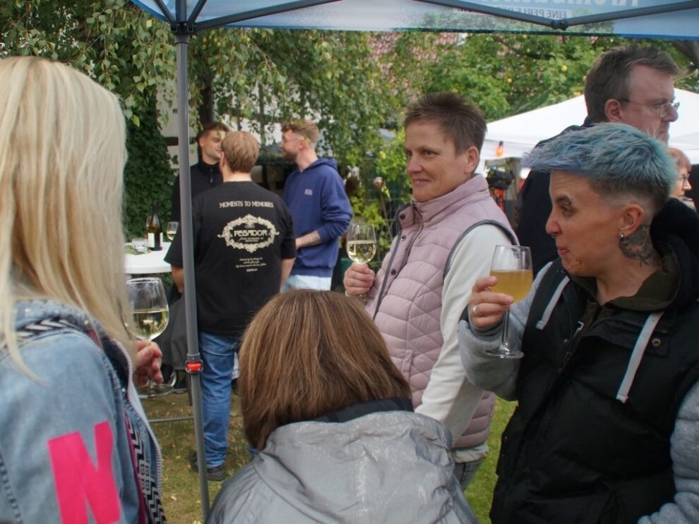 Mehrere Gäste stehen mit Wein und einem hellen Getränk unter einem Pavillon.