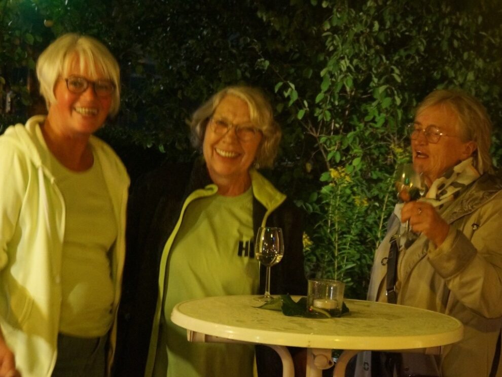 Drei Frauen lächeln am Abend mit Weingläsern an einem Stehtisch.