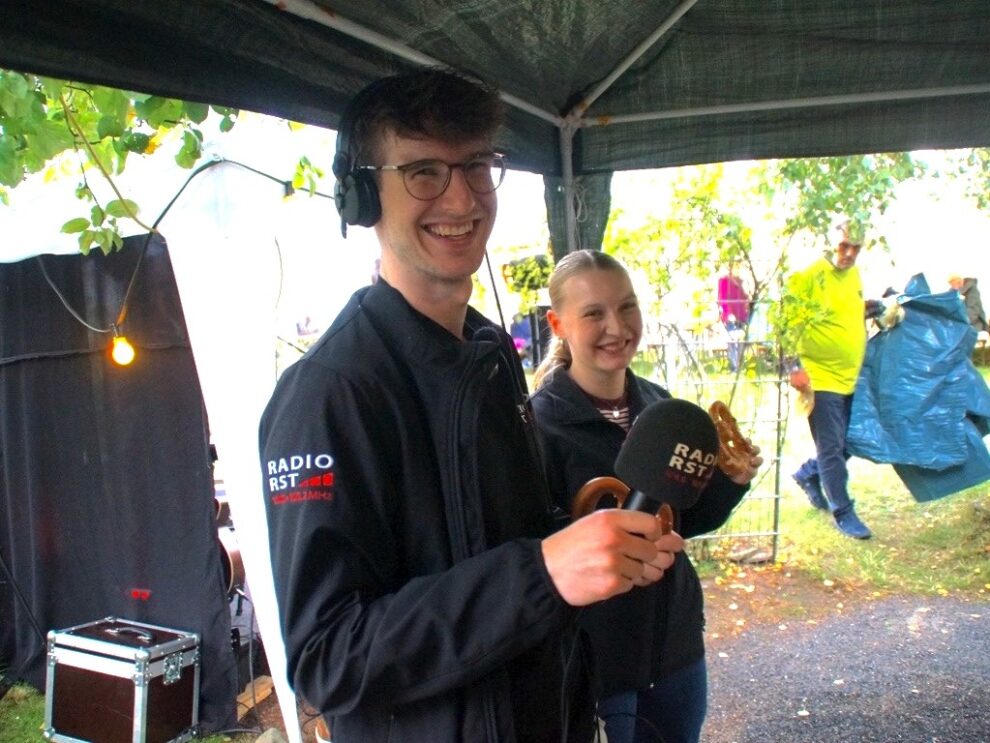 Ein Radioreporter und eine junge Frau lachen mit Mikrofon und Brezel in die Kamera.