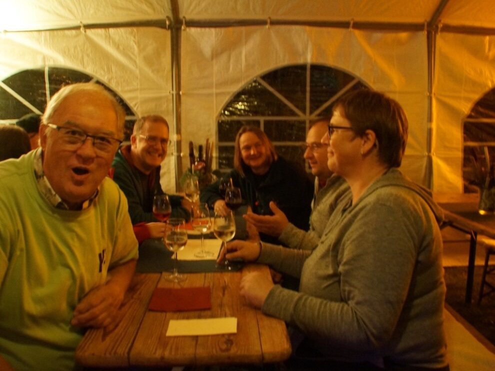 Mehrere Gäste sitzen am Abend mit Weingläsern an einer langen Tafel im Festzelt.
