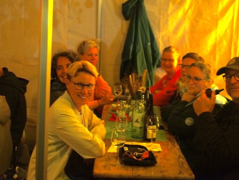 Eine größere Gruppe lächelt am Abend an einer Tafel mit Weinflaschen und Gläsern in die Kamera.