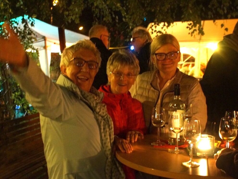 Drei gut gelaunte Frauen stehen abends mit Weinflasche und Gläsern an einem beleuchteten Stehtisch.